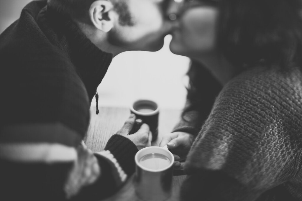 Bildet viser et par som sitter med hver sin kaffekopp og kysser.