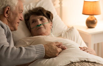 Trøst fra pårørende Bildet viser en syk eldre dame som ligger i senga med neseslange. En eldre mann holder henne i hånda og kikker på henne
