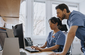 Sykepleiere foran PC Bildet viser en kvinnelig sykepleier som sitter foran en PC. En mannlig sykepleier står bak henne og kikker på skjermen